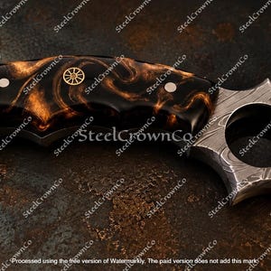 Puede incluir: Un cuchillo curvo con un mango de patr&oacute;n jaspeado en negro y cobre, asegurado por dos remaches plateados y un emblema central de rueda dorada. La hoja muestra un patr&oacute;n distintivo de acero de Damasco con un orificio circular para el dedo.