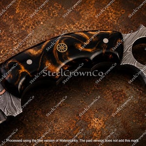 Puede incluir: Un cuchillo con hoja de acero Damasco que presenta una incrustaci&oacute;n de cobre ondulada. El mango es de resina veteada en negro y &aacute;mbar, fijado con dos pasadores plateados y un emblema de rueda dorado. Tiene un pomo en forma de anillo estrellado.