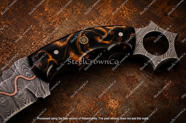 Puede incluir: Un cuchillo con hoja de acero Damasco que presenta una incrustaci&oacute;n de cobre ondulada. El mango es de resina veteada en negro y &aacute;mbar, fijado con dos pasadores plateados y un emblema de rueda dorado. Tiene un pomo en forma de anillo estrellado.