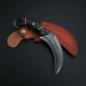 Puede incluir: Un cuchillo karambit curvo con hoja de acero damasco estampada y mango negro con pasadores de lat&oacute;n y un anillo para el dedo. El cuchillo descansa sobre una funda de cuero marr&oacute;n con costuras decorativas y un cierre de bot&oacute;n met&aacute;lico.