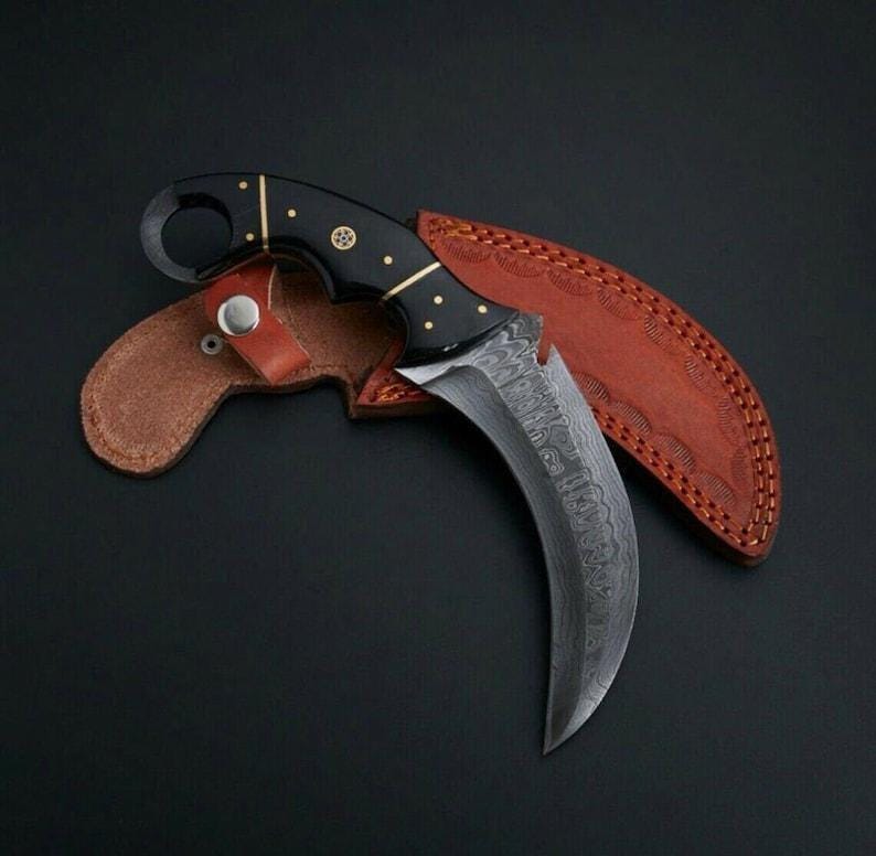 Puede incluir: Un cuchillo karambit curvo con hoja de acero damasco estampada y mango negro con pasadores de lat&oacute;n y un anillo para el dedo. El cuchillo descansa sobre una funda de cuero marr&oacute;n con costuras decorativas y un cierre de bot&oacute;n met&aacute;lico.
