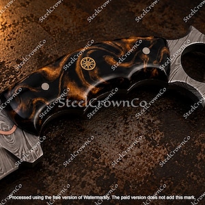 Puede incluir: Un cuchillo con hoja de acero de Damasco que presenta una incrustaci&oacute;n de cobre ondulada y un mango jaspeado en negro y &aacute;mbar. El mango incluye un emblema circular de color dorado y un pomo en forma de estrella con un orificio central.