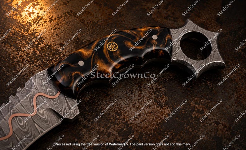 Puede incluir: Un cuchillo con hoja de acero de Damasco que presenta una incrustaci&oacute;n de cobre ondulada y un mango jaspeado en negro y &aacute;mbar. El mango incluye un emblema circular de color dorado y un pomo en forma de estrella con un orificio central.