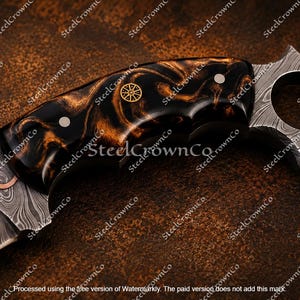 Puede incluir: Un cuchillo con hoja de acero de Damasco con una incrustaci&oacute;n de cobre ondulada. El mango es de resina veteada negra y &aacute;mbar con dos pasadores plateados y un emblema circular dorado. El pomo es un anillo en forma de estrella de acero de Damasco.