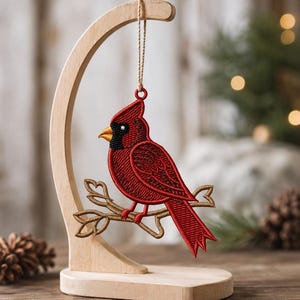May include: A red cardinal bird ornament with black facial details and a yellow beak, embroidered on a tan branch. The ornament hangs from a light-colored wooden stand with a curved arm and a flat base, suspended by a thin twine string.