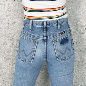 Puede incluir: Vaqueros Wrangler de mezclilla azul claro con cintura alta y dise&ntilde;o cl&aacute;sico de bolsillo trasero. Los vaqueros presentan el logotipo de Wrangler en el bolsillo trasero y se combinan con una camiseta a rayas con colores del arco&iacute;ris.