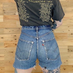 Könnte beinhalten: Eine blaue Jeans-Shorts mit ausgefransten Kanten. Die Shorts haben einen hohen Bund und sind kurz geschnitten, enden oberhalb der Knie. Die Shorts haben ein klassisches Fünf-Taschen-Design und einen roten Streifen an der rechten Gesäßtasche.