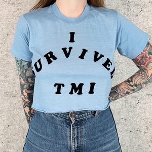 May include: Light blue crop top with the text "I SURVIVED TMI" in black. The shirt has a raw hem and short sleeves. The model is wearing blue jeans.