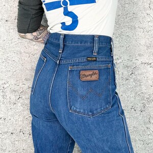 May include: Blue denim Wrangler jeans with a brown leather patch on the back pocket. The jeans have a high waist and a classic fit. The person is wearing a white t-shirt with blue text that says "BLUE OF CULT" and gray sleeves.