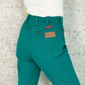 May include: Teal-colored denim jeans with a high waist and a brown leather patch on the back pocket. The jeans have a classic five-pocket design and a red tag. The person is wearing a white top.