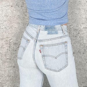May include: Light wash denim jeans with a high waist and classic five-pocket design. The jeans feature a red tag on the back pocket and a blue patch at the waistband. The model is wearing a light blue top.