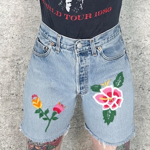 Könnte beinhalten: Hellblaue Jeansshorts mit gestickten Blumenmustern in Rosa, Gelb und Rot. Die Shorts haben einen ausgefransten Saum und einen Knopfverschluss. Das Model trägt ein schwarzes T-Shirt mit dem Text "WORLD TOUR 1980".