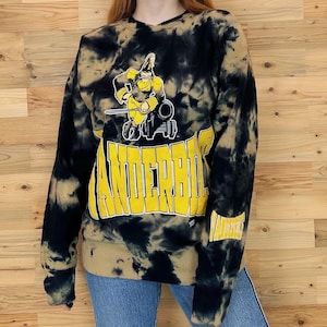 May include: Black and yellow tie-dye sweatshirt with a cartoon mascot wearing a hat and holding a cannon. The text "Vanderbilt" is printed in yellow on the front of the sweatshirt.
