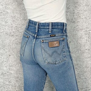 May include: Light blue denim jeans with a brown leather Wrangler patch on the back pocket. The jeans have a classic five-pocket design and a high waist. The jeans are worn with a white top.