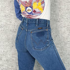 K&ouml;nnte beinhalten: Blaue Wrangler Jeans mit hohem Bund und klassischem R&uuml;ckentaschendesign. Die Jeans haben einen geraden Beinverlauf und das Wrangler-Logo auf der Ges&auml;&szlig;tasche. Das Model tr&auml;gt ein lang&auml;rmliges Shirt mit einem grafischen Design.