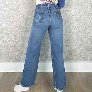 K&ouml;nnte beinhalten: Hellblaue Jeans mit hoher Taille und weitem Bein. Die Jeans haben ein klassisches F&uuml;nf-Taschen-Design und einen braunen Lederaufn&auml;her auf der R&uuml;ckseite. Die Person tr&auml;gt ein wei&szlig;es und blaues Langarmshirt mit dem Text "Bite the Dust Tour".