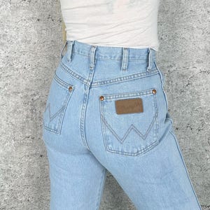 May include: Pale blue denim jeans with a classic fit. Features include a brown leather patch with the "Wrangler" logo on the back pocket, copper rivets, and a high waist. The jeans have a traditional five-pocket design and a straight leg.