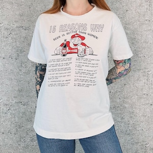 以下が含まれることがあります： 「15 REASONS WHY BEER IS BETTER THAN WOMEN」の文字がプリントされたオフホワイトのTシャツ。ビール瓶とジョッキを持った男性の漫画のイラストが描かれています。半袖でクルーネックのシャツです。