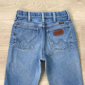 Op de afbeelding: Lichtblauwe spijkerbroek met een klassiek ontwerp met vijf zakken. De jeans heeft een bruine leren patch met het "Wrangler"-logo op de achterzak. De achterzakken hebben een kenmerkende "W"-stitching.