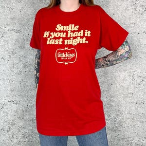 以下が含まれることがあります： 「Smile if you had it last night.」とLittle Kings Cream Aleのロゴが書かれた白いグラフィックがプリントされた赤いTシャツです。