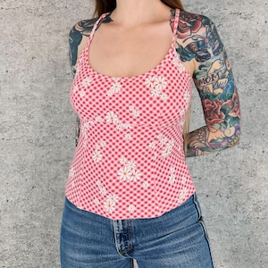 May include: A pink and white gingham print camisole top with white daisy floral accents. The top has thin straps and a scoop neckline. The model is wearing blue jeans.