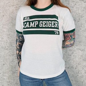 Könnte beinhalten: Weißes T-Shirt mit grünen Zierleisten und dem Text "BSA CAMP GEIGER '78". Das Shirt hat ein grünes Band über der Brust. Das Shirt ist im Vintage-Stil.