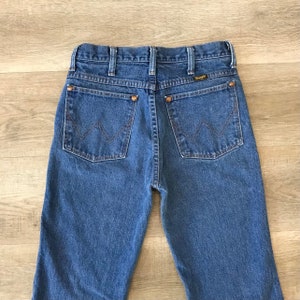 K&ouml;nnte beinhalten: Eine blaue Jeanshose mit klassischer gerader Passform. Die Jeans hat einen braunen Leder-Patch mit dem Wrangler-Logo auf dem hinteren Bund. Die Ges&auml;&szlig;taschen haben eine charakteristische W-f&ouml;rmige Steppnaht.