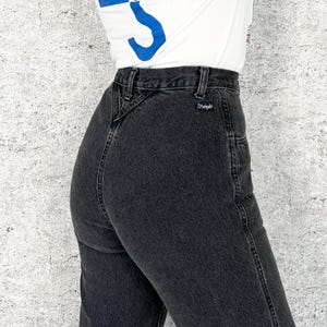 Pu&ograve; includere: Primo piano di jeans neri. I jeans hanno una vita alta e un classico design a cinque tasche. Il logo Wrangler &egrave; visibile sulla tasca posteriore. I jeans sono indossati con una maglietta bianca con una grafica blu.