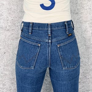 May include: Close-up of the back of blue denim jeans with the Wrangler logo on the back pocket. The jeans have a classic five-pocket design and a high waist. The person is wearing a cream-colored shirt with a blue design.