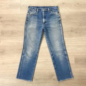 May include: Light blue denim jeans with a classic five-pocket design. The jeans have a straight leg cut with a raw, unfinished hem. The denim shows signs of wear and fading, giving them a vintage look. The jeans are laid flat on a light wood surface.