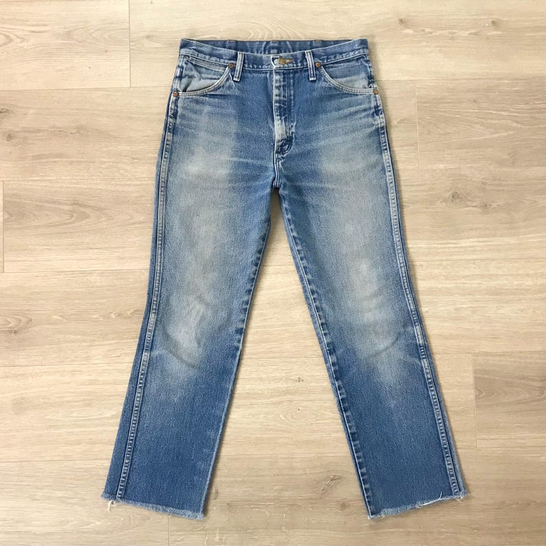May include: Light blue denim jeans with a classic five-pocket design. The jeans have a straight leg cut with a raw, unfinished hem. The denim shows signs of wear and fading, giving them a vintage look. The jeans are laid flat on a light wood surface.