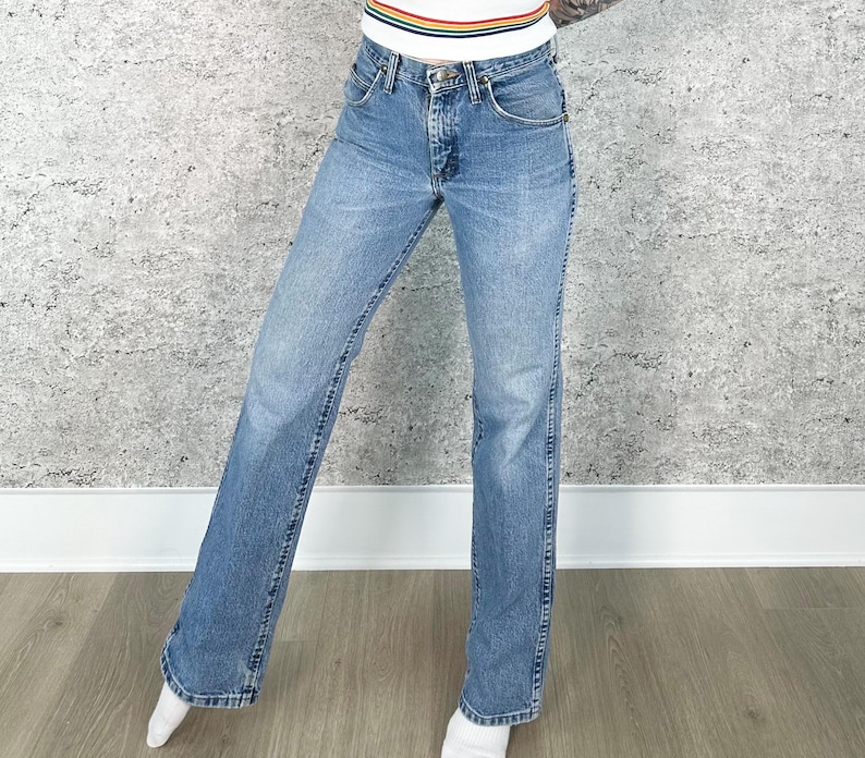 Puede incluir: Vaqueros de mezclilla azul claro con un dise&ntilde;o cl&aacute;sico de cinco bolsillos. Los vaqueros tienen un corte de pierna recta y un lavado descolorido. El modelo lleva una camiseta blanca con un dise&ntilde;o de rayas arco&iacute;ris.