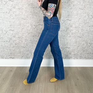 May include: Dark blue denim jeans with a flared leg. The jeans have a classic five-pocket design and a high waist. The denim is a medium wash, and the jeans appear to be vintage. The jeans are worn with a black top.