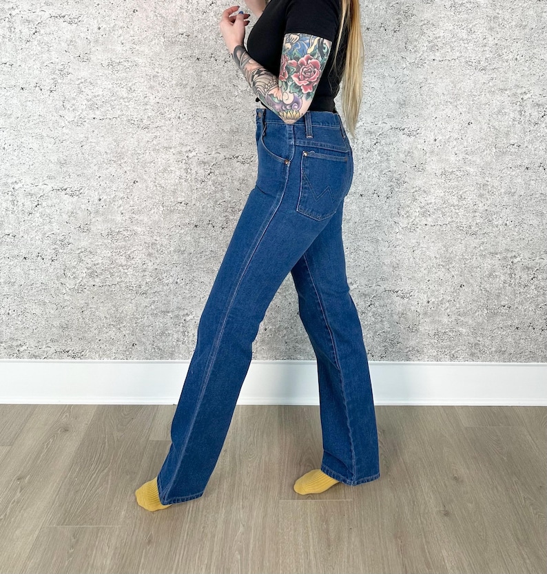May include: Dark blue denim jeans with a flared leg. The jeans have a classic five-pocket design and a high waist. The denim is a medium wash, and the jeans appear to be vintage. The jeans are worn with a black top.