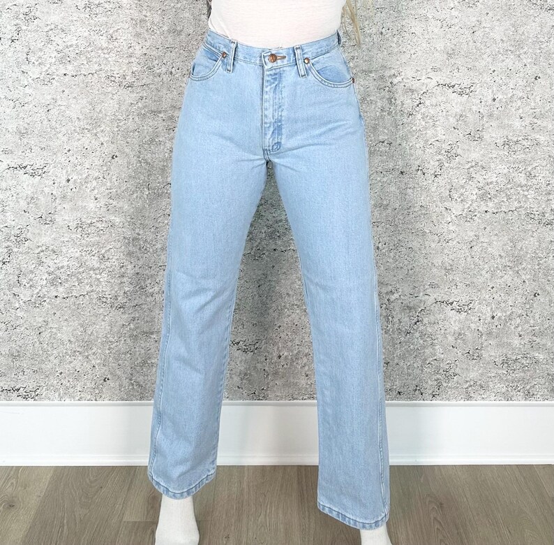 May include: Pale blue denim jeans with a classic five-pocket design. The jeans feature a high waist and a straight leg fit. The denim is a light wash, and the jeans have a button and zip closure.