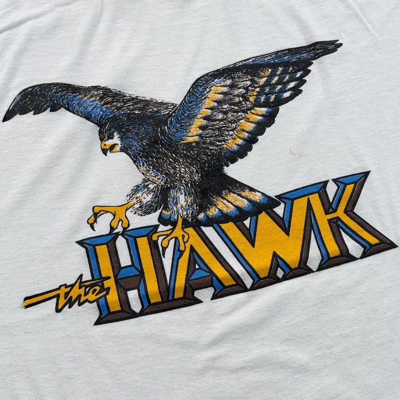 May include: White t-shirt featuring a detailed illustration of a hawk with outstretched wings. The hawk is depicted in shades of black, blue, and yellow, perched above the text "the HAWK" in a stylized font. The design is centered on the shirt.