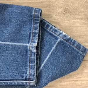 May include: A pair of blue denim jeans featuring a medium wash with visible fading and whiskering patterns. The hem of the trouser legs shows signs of wear with slight fraying along the bottom edges and thick, contrasting copper-coloured stitching.