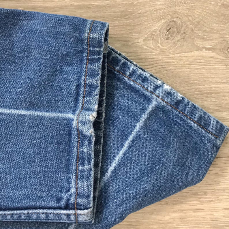 May include: A pair of blue denim jeans featuring a medium wash with visible fading and whiskering patterns. The hem of the trouser legs shows signs of wear with slight fraying along the bottom edges and thick, contrasting copper-coloured stitching.