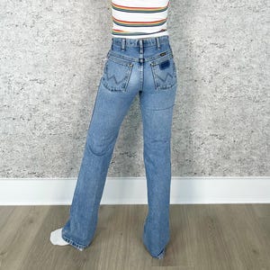 Puede incluir: Vaqueros Wrangler de mezclilla azul claro con pernera acampanada. Los vaqueros tienen un estilo cl&aacute;sico de cinco bolsillos y un parche con el logo de Wrangler en el bolsillo trasero. La persona tambi&eacute;n lleva una camiseta a rayas.