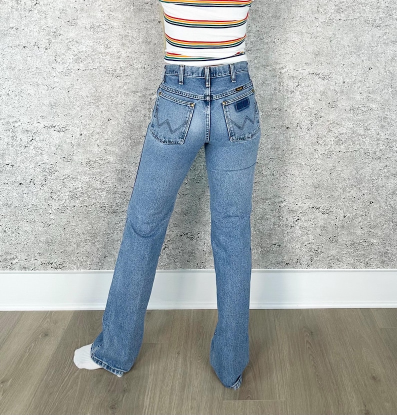 Puede incluir: Vaqueros Wrangler de mezclilla azul claro con pernera acampanada. Los vaqueros tienen un estilo cl&aacute;sico de cinco bolsillos y un parche con el logo de Wrangler en el bolsillo trasero. La persona tambi&eacute;n lleva una camiseta a rayas.