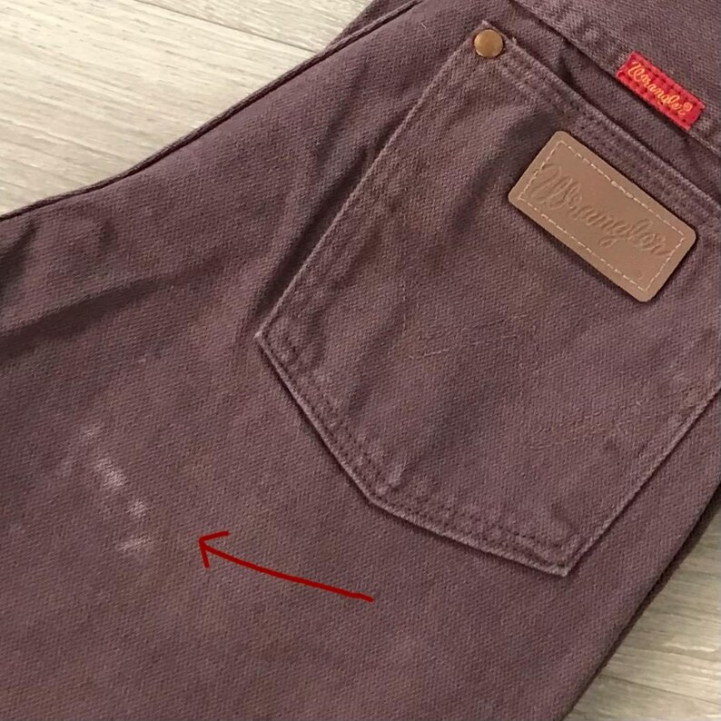 Op de afbeelding: Close-up van een paar stoffig roze Wrangler jeans. De jeans hebben een achterzak met een bruine leren Wrangler patch en een rood Wrangler label. De stof vertoont wat slijtage, met een paar witte vlekken aan de linkerkant.