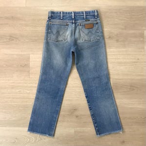 May include: Light blue denim jeans with a classic five-pocket design. The jeans have a faded wash, a brown leather patch, and the Wrangler logo on the back pockets. The hem is frayed.