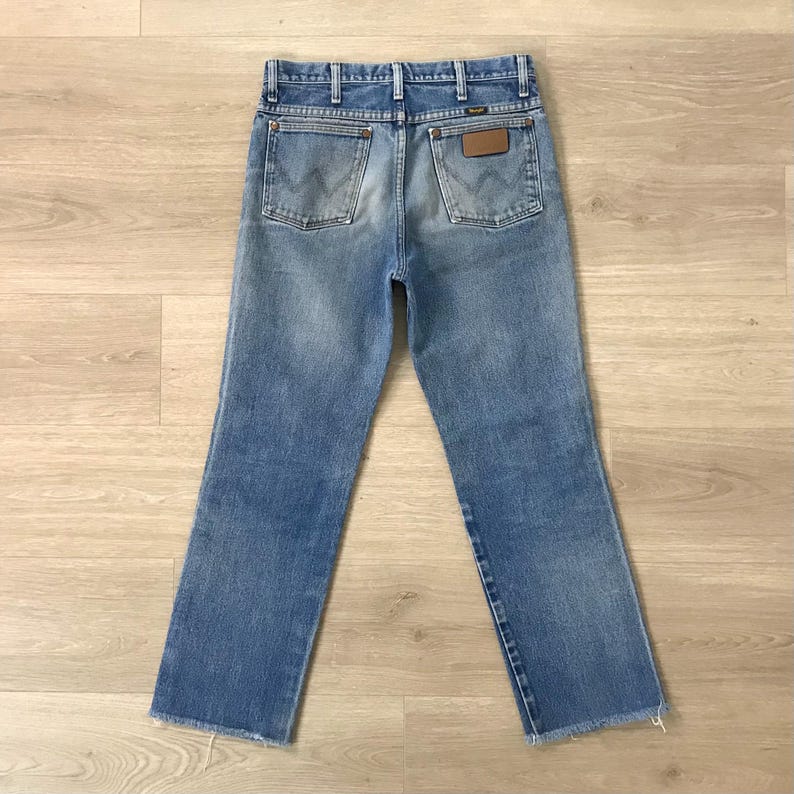 May include: Light blue denim jeans with a classic five-pocket design. The jeans have a faded wash, a brown leather patch, and the Wrangler logo on the back pockets. The hem is frayed.
