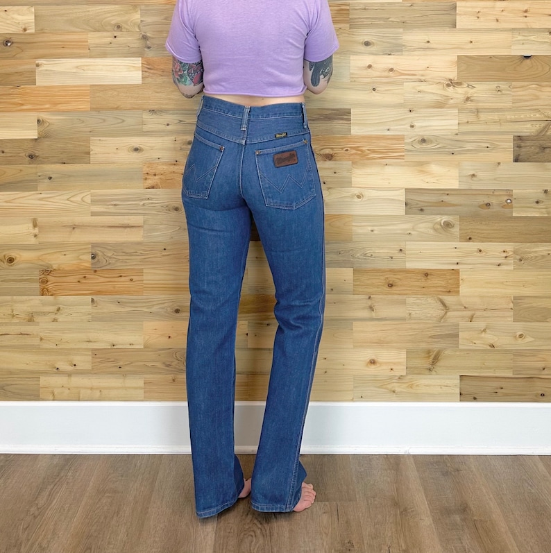 Pu&ograve; includere: Jeans Wrangler in denim blu con vita alta e gamba svasata. I jeans hanno una toppa in pelle marrone sulla tasca posteriore. La persona indossa un top corto viola.