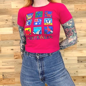 Op de afbeelding: Een felroze T-shirt met korte mouwen voorzien van een raster van negen kleurrijke illustraties van Australische dieren en de Australische vlag. Het woord AUSTRALIA staat in grote, veelkleurige letters onderaan het grafische ontwerp.