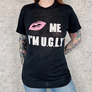 May include: A black short-sleeve t-shirt featuring a graphic of pink lips and white text that reads "ME" above the word "I'M U.G.L.Y." The shirt has a crew neckline and a lightweight, slightly sheer fabric construction with a standard fit.