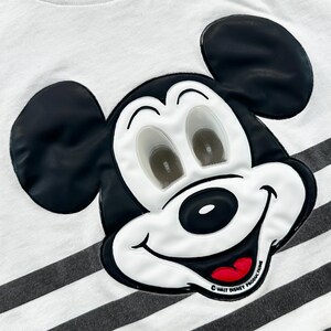 May include: Close-up of a white t-shirt with black and gray horizontal stripes. The shirt features a large, raised Mickey Mouse face in black, white, and red. The text "&copy; WALT DISNEY PRODUCTIONS" is printed on the bottom right.