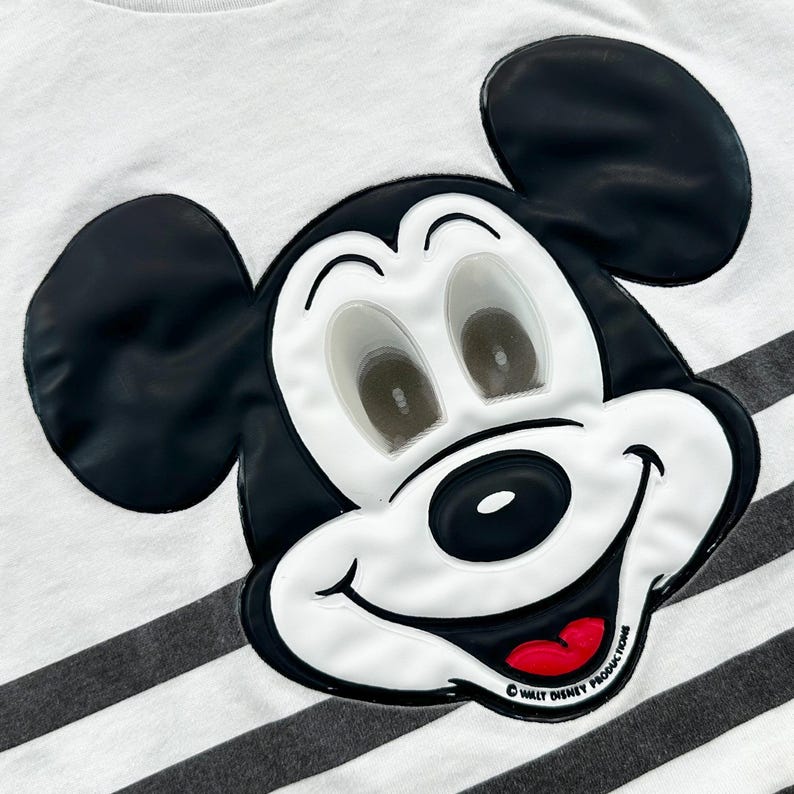 May include: Close-up of a white t-shirt with black and gray horizontal stripes. The shirt features a large, raised Mickey Mouse face in black, white, and red. The text "&copy; WALT DISNEY PRODUCTIONS" is printed on the bottom right.