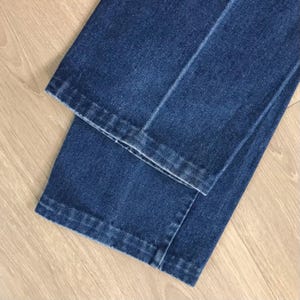 May include: Close-up of folded blue denim jeans. The jeans are a medium wash with visible seams and a classic straight leg cut. The fabric appears to be sturdy and well-constructed, with a slight sheen.