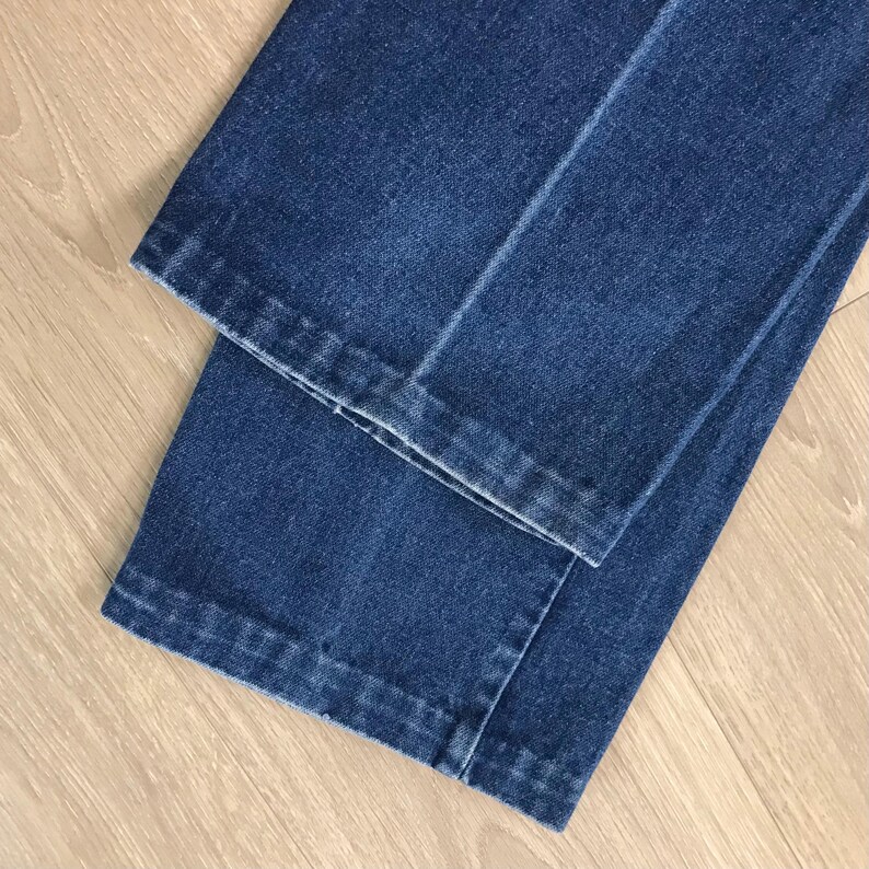 May include: Close-up of folded blue denim jeans. The jeans are a medium wash with visible seams and a classic straight leg cut. The fabric appears to be sturdy and well-constructed, with a slight sheen.